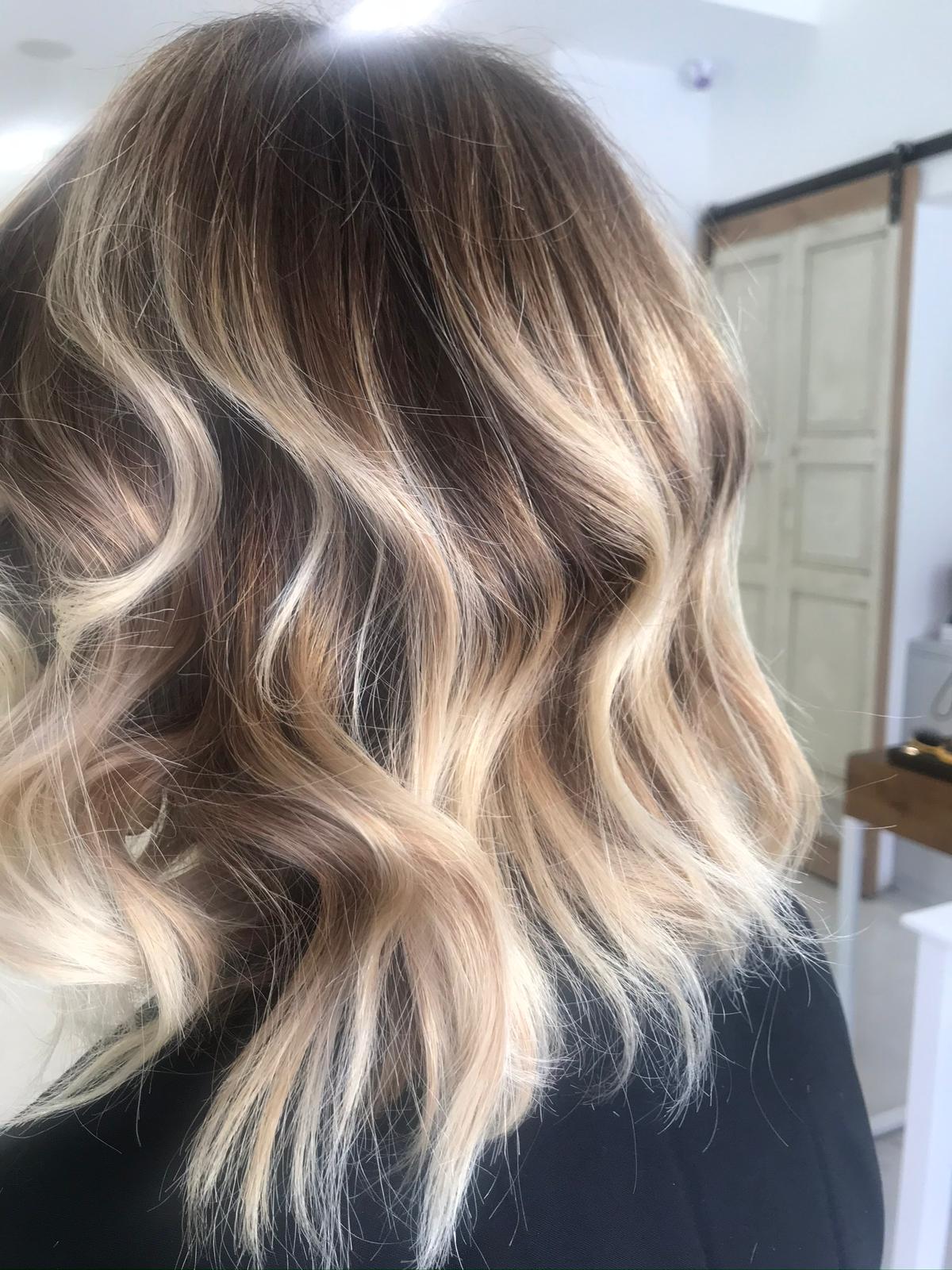 Capelli mossi con balayage dalla radice castana alle punte biondo miele, su giacca nera, in salone con porta in legno chiaro