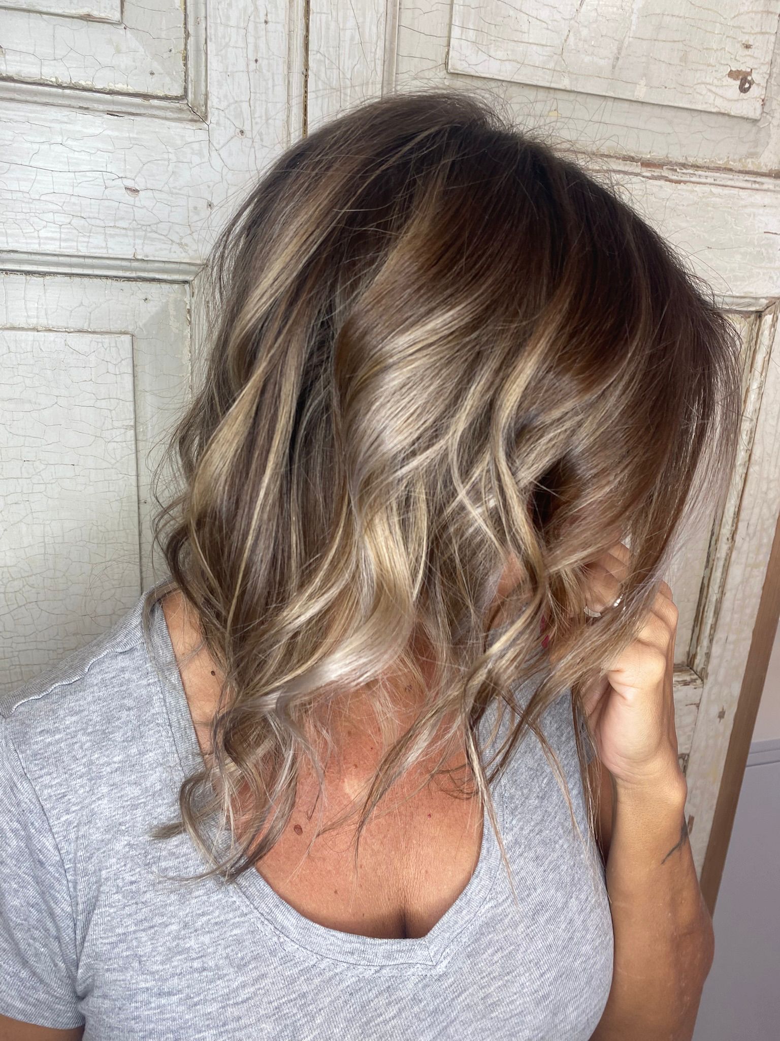 Capelli castani mossi di media lunghezza con shatush biondo cenere, davanti a una vecchia porta bianca screpolata