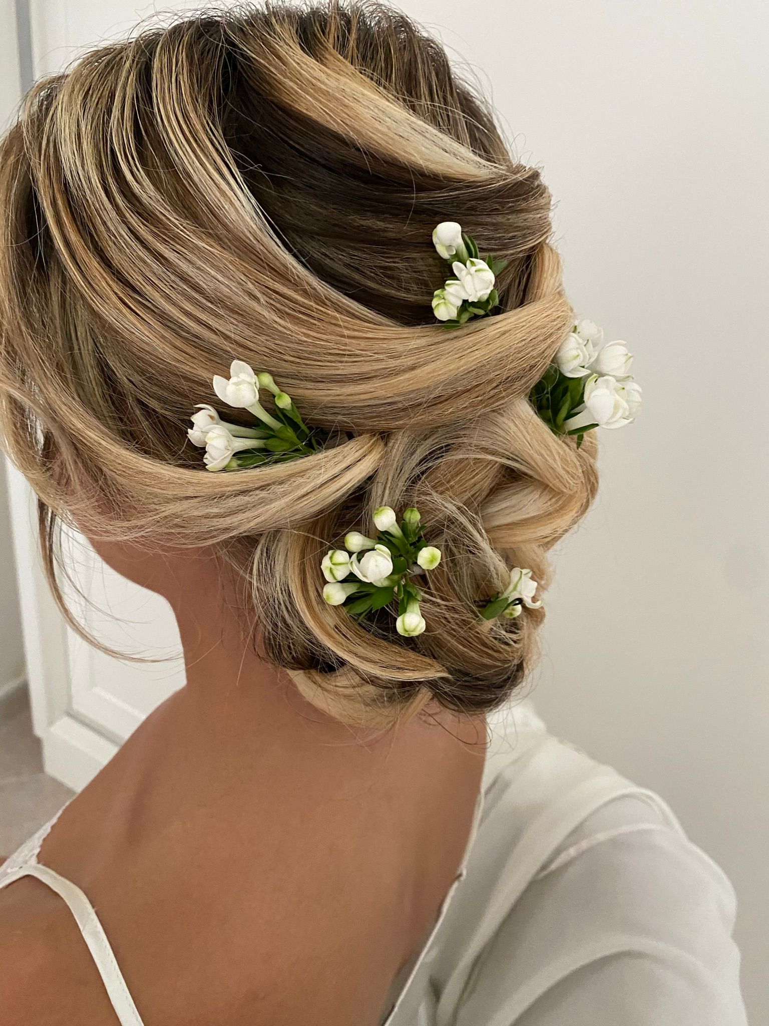 Raccolto morbido su capelli biondi con mèches, decorato con boccioli di fiori bianchi, visto da dietro su spalle scoperte